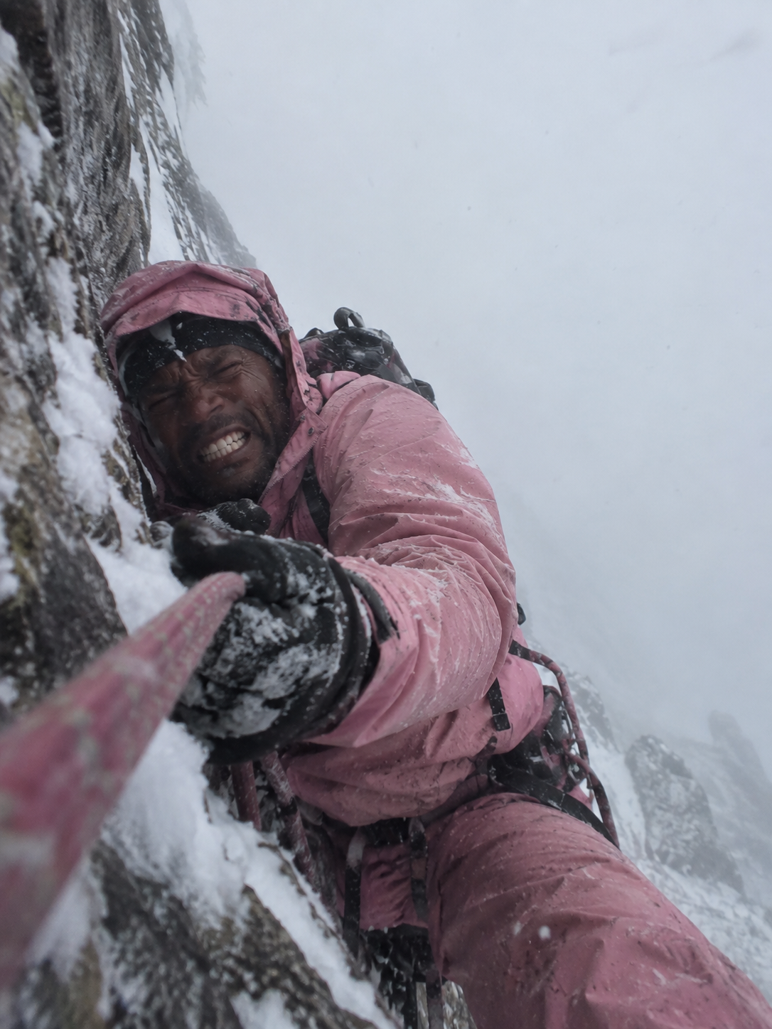 A climber in a pink snowsuit pulling themselves up a sheer rock face in a snowstorm